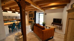 A living room with a wooden ceiling, a sofa, a television, and a kitchen in the background. - The Munster valley stable (Breitenbach-Haut-Rhin)