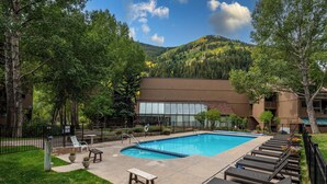 A swimming pool with lounge chairs, a picnic table, and a building with large windows in the background. - Mountain Retreat in East Vail, 2BR/2BA Condo w/ Pool & Hot Tub (Vail)