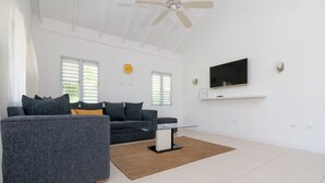 A modern living room with a grey sofa, a glass coffee table, and a flat-screen TV mounted on the wall. - Eshae Living - Uniquely Modern Beach House 3 (Plantation Village)