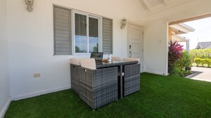 An outdoor seating area with a wicker table and chairs, a laptop on the table, and a door in the background. - Eshae Living - Uniquely Modern Beach House 3 (Plantation Village)