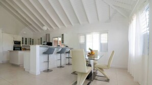 A spacious kitchen with white cabinetry, a central island with bar stools, and a dining area with a glass table and chairs. - Eshae Living - Uniquely Modern Beach House 3 (Plantation Village)