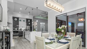 A modern kitchen with white cabinets, stainless steel appliances, and a dining area with a glass table set for four. - Golden Acres Sports Ranch – Luxe Retreat with Pickleball, Pool & Games (Naples)