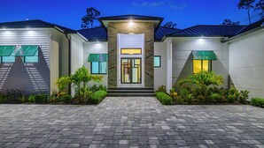 A modern two-story house with a paved driveway, a central entrance, and green shutters on the windows. - Golden Acres Sports Ranch – Luxe Retreat with Pickleball, Pool & Games (Naples)