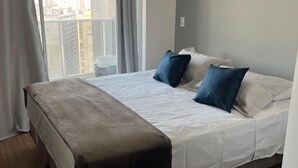 A bedroom with a bed, a bench, a window with curtains, and a view of buildings. - Estúdio aconchegante na Sé (São Paulo)