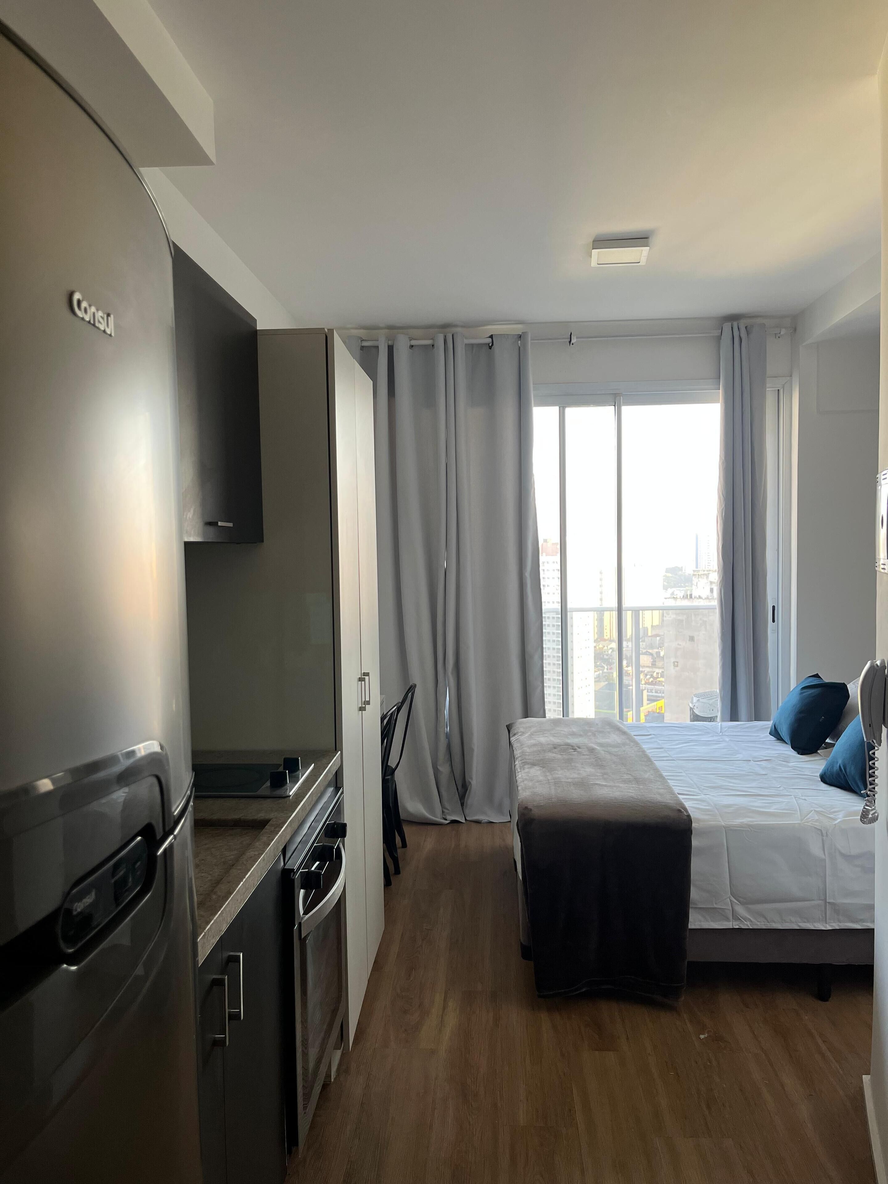 Ein modernes Hotelzimmer mit einer Küchenzeile, einem Bett mit dunklem Kopfteil und einem großen Fenster mit Vorhängen.