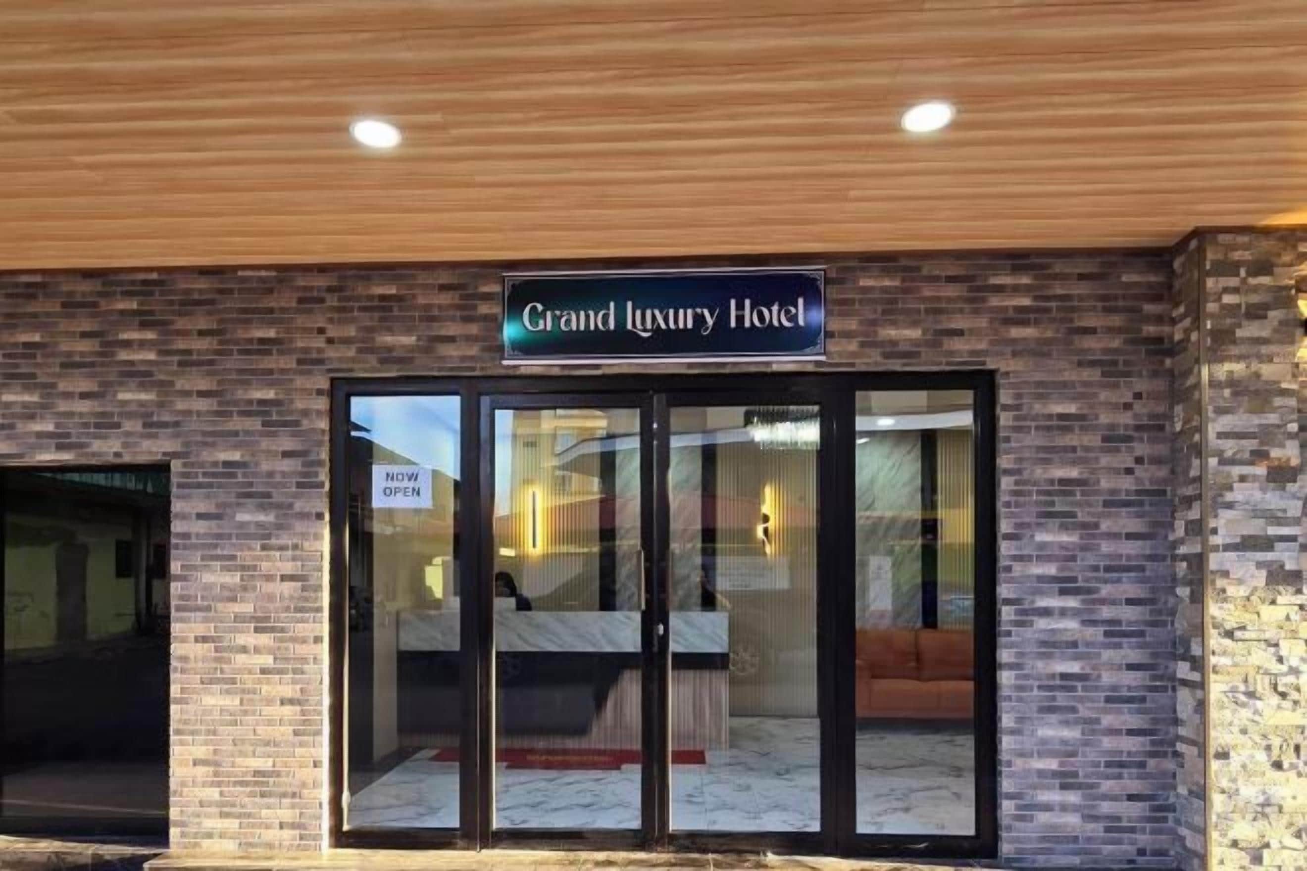 The exterior of the Grand Luxury Hotel with a brick facade, a glass entrance, and a sign indicating the hotel is now open.