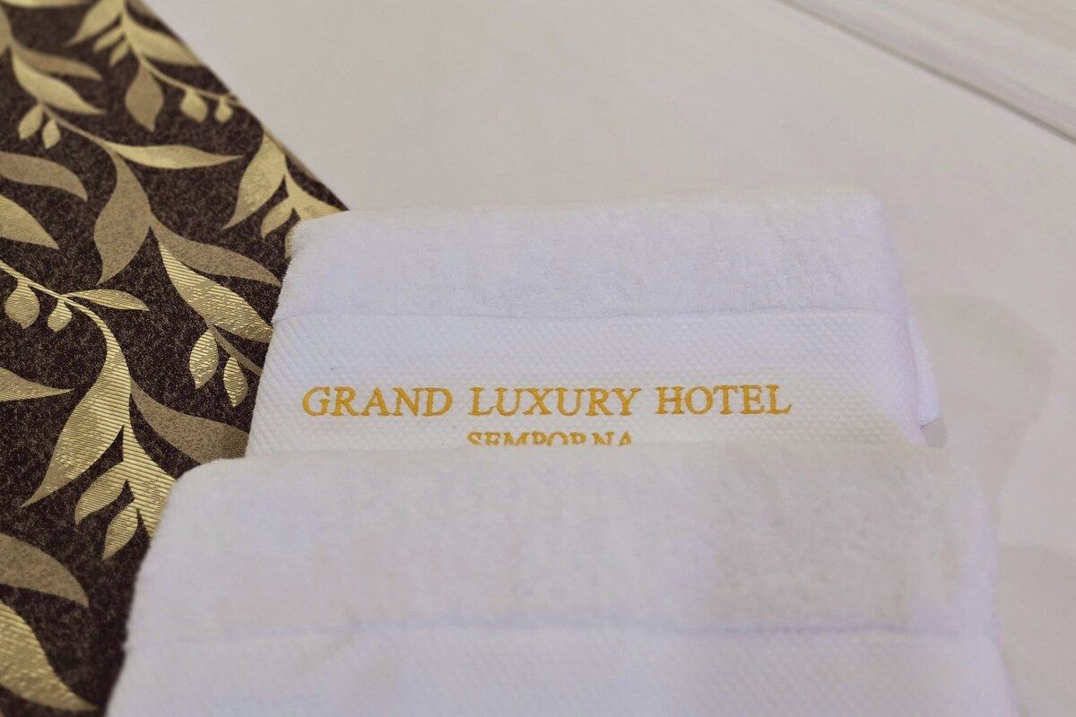 Un lit d’hôtel avec une couvre-lit blanche et un coussin décoratif. Une serviette blanche portant l’inscription « GRAND LUXURY HOTEL » est posée sur le lit.