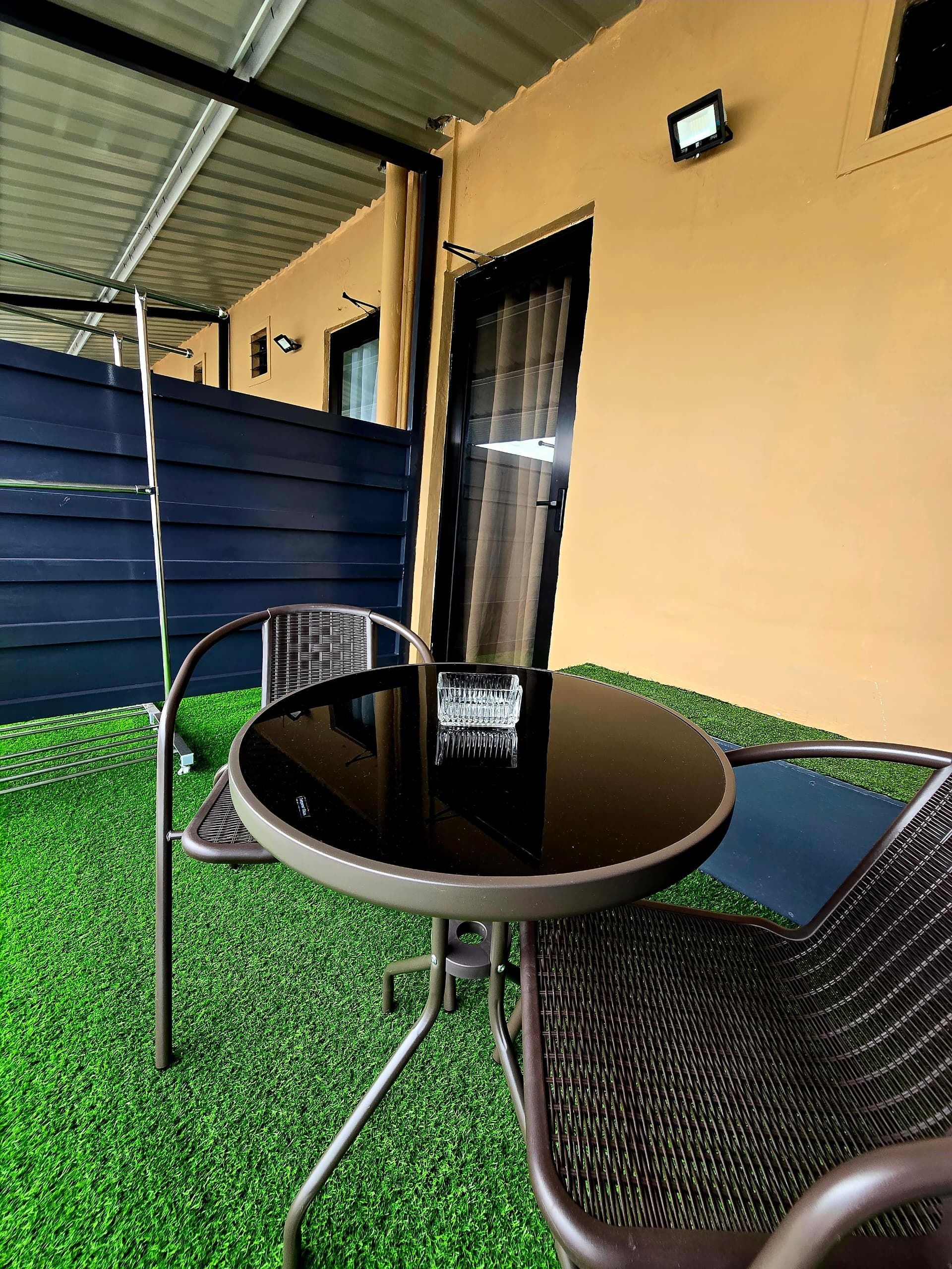 An outdoor seating area with a round table and two chairs, a metal fence, and a building in the background.