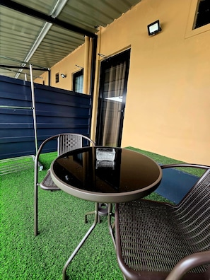 Un área de estar al aire libre con una mesa redonda y dos sillas, una cerca de metal y un edificio al fondo.