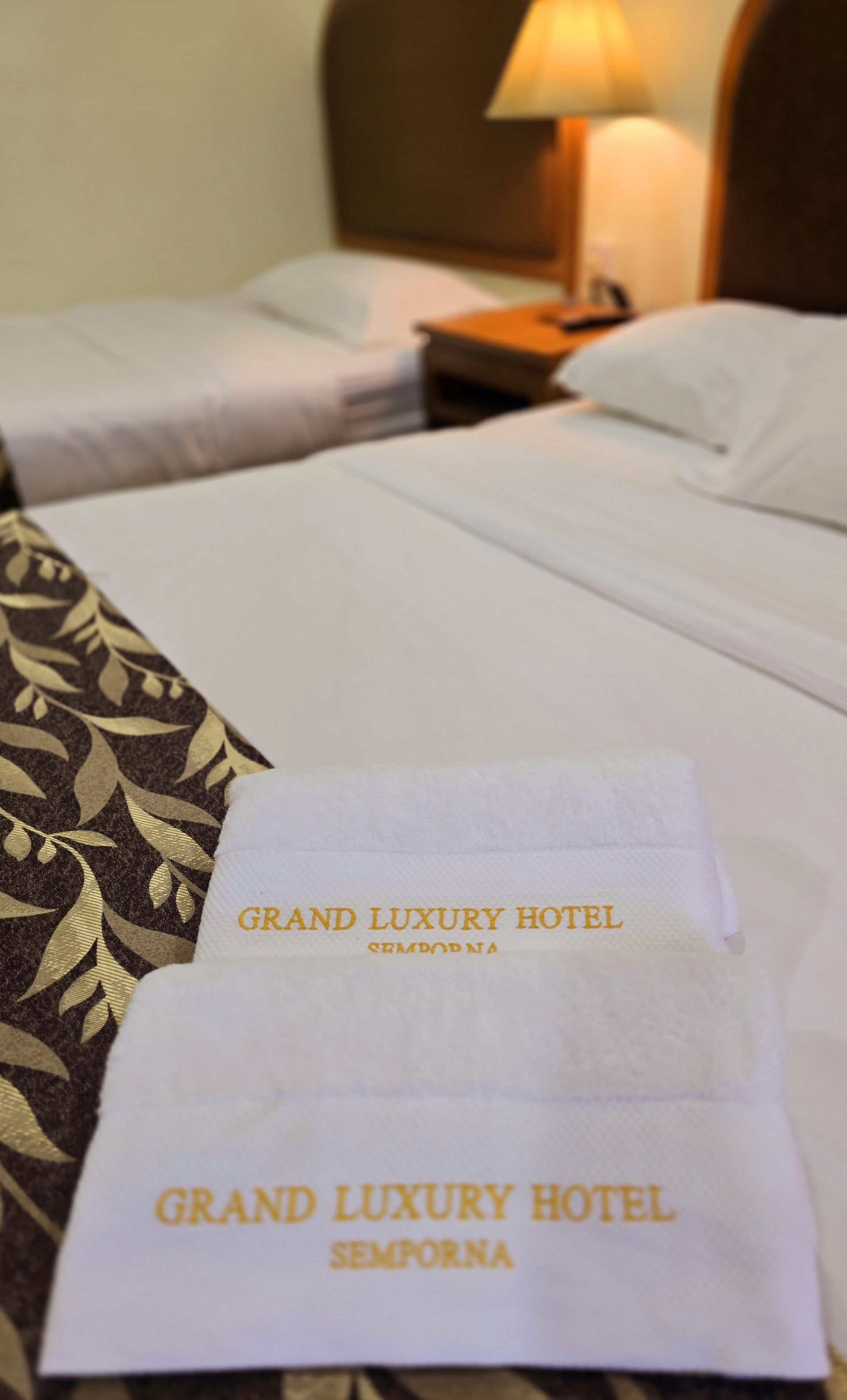 A hotel bed with a white bedspread and a decorative pillow. A white towel with 'GRAND LUXURY HOTEL' text is placed on the bed.