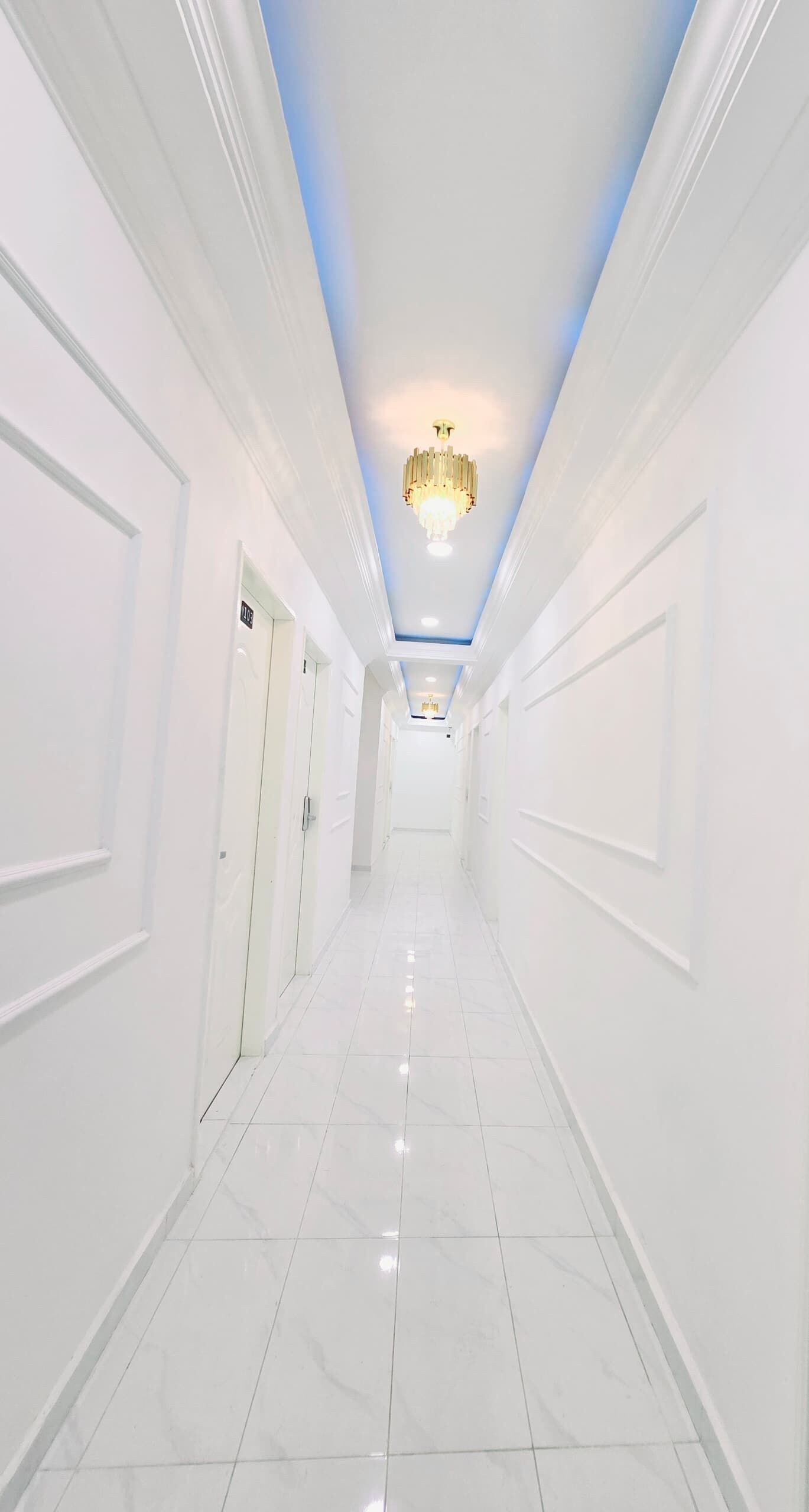 A long, narrow hallway with white walls and a tiled floor, leading to a room with a door at the end.