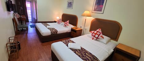 A hotel room with two beds, wooden flooring, a desk, a chair, a lamp, and a TV.
