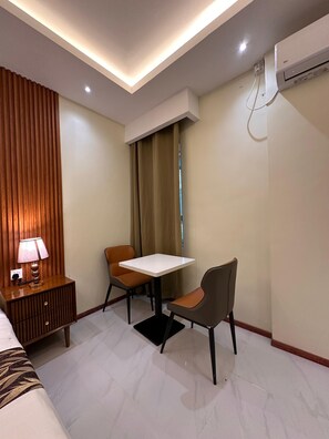 Habitación de hotel con una cama, una mesita de noche con lámpara, una pequeña mesa con dos sillas, un equipo de aire acondicionado y una ventana con cortinas.