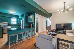 Une cuisine moderne avec des meubles de couleur verte, un coin bar avec des tabourets, un espace repas avec un téléviseur et un salon avec un canapé et une table basse.