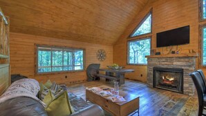 A log cabin interior with a fireplace, a sofa, a dining table, and large windows. - Contemporary Cabin w/ Hot Tub - 2BR, 2BA, Sleeps 8 (Blue Ridge)