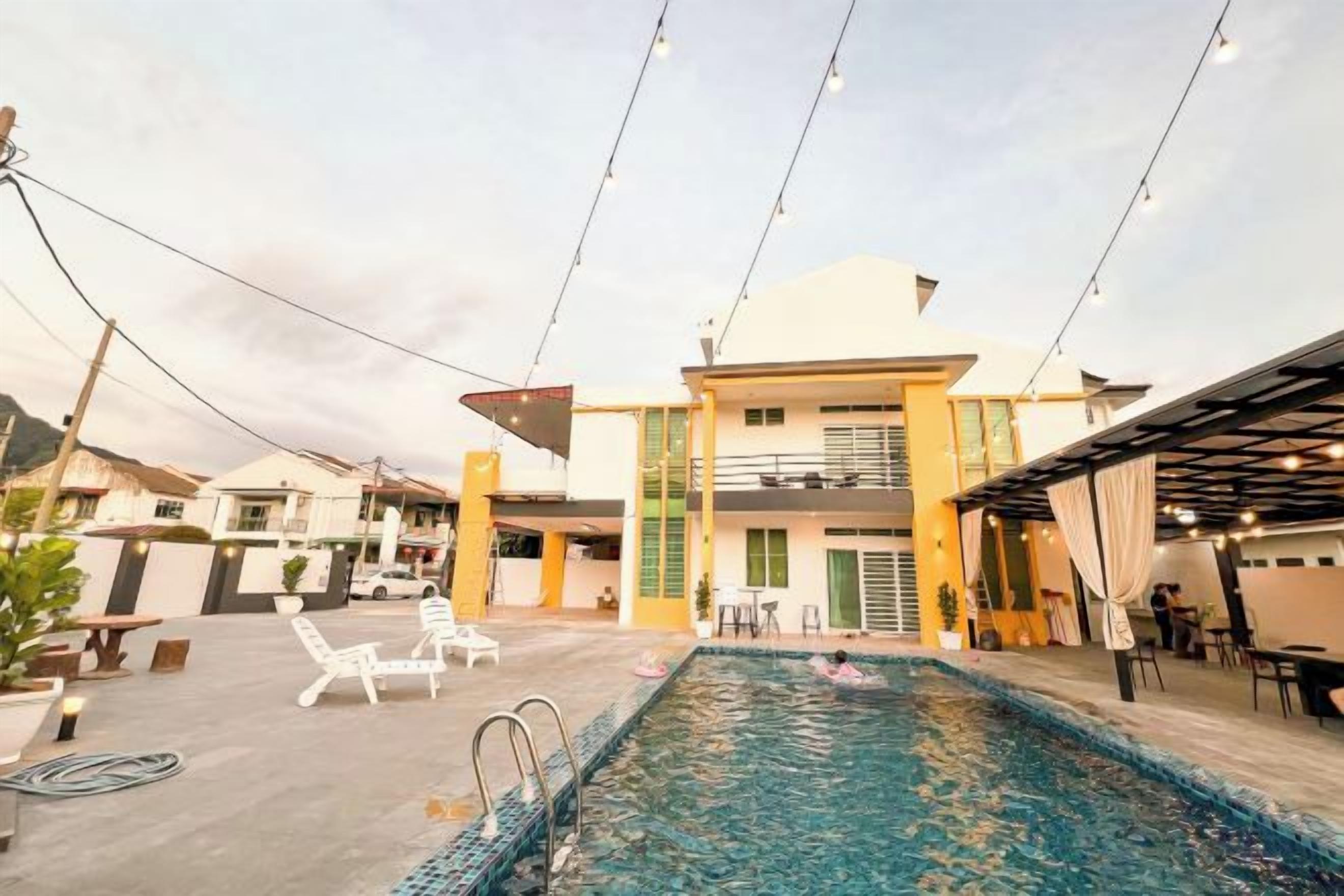 Una piscina con sillas de descanso, una zona de estar techada y un edificio de techo plano con varias ventanas.