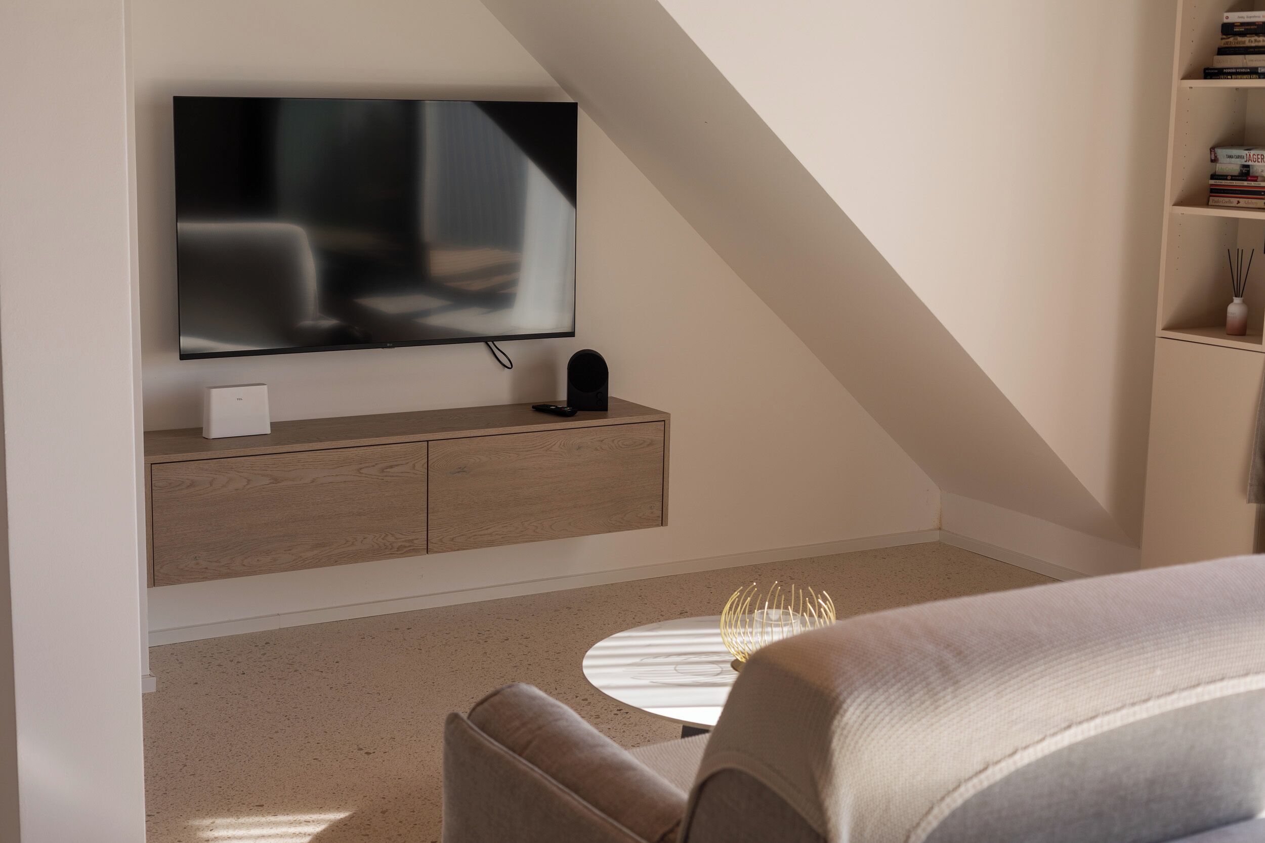 Ein modernes Schlafzimmer mit an der Wand montiertem Fernseher, einem Nachttisch, einem runden Couchtisch und einem Bücherregal.