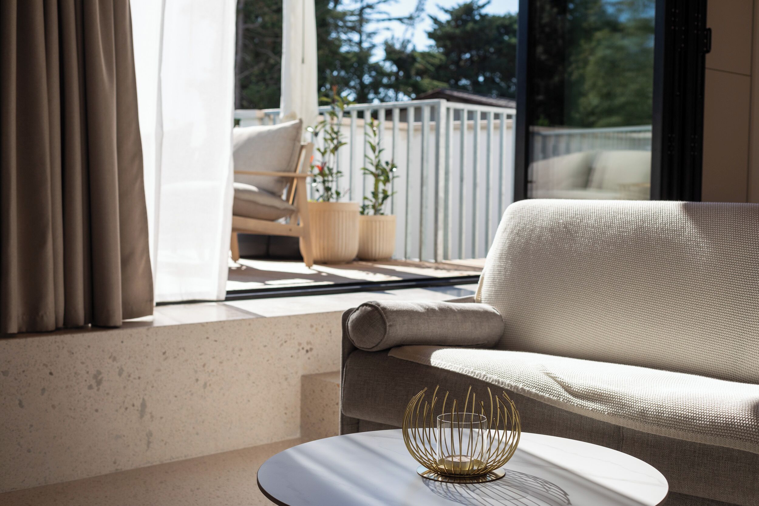A modern living room with a sofa, a round table, and a view of a balcony with plants and a railing.