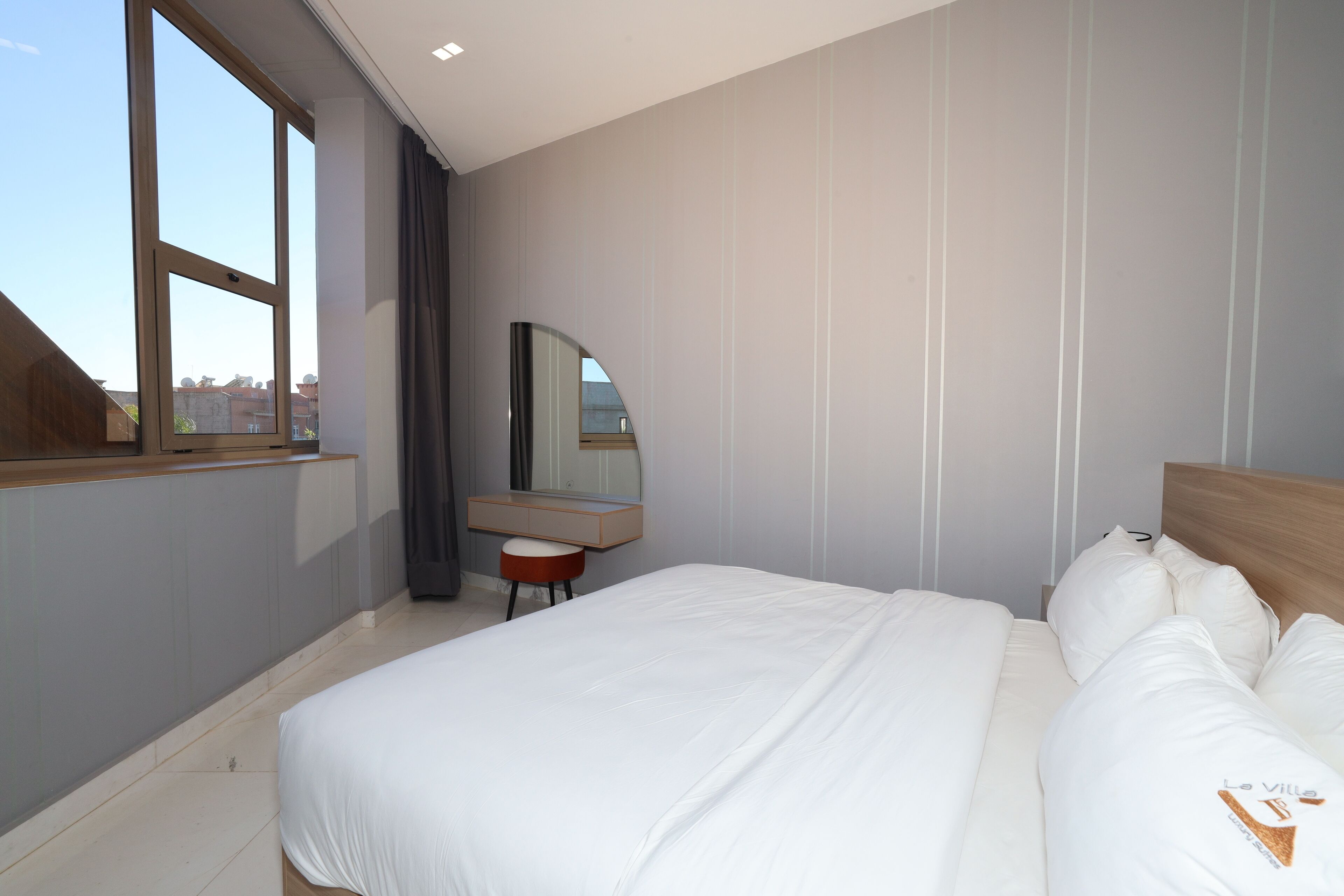 Une chambre d’hôtel moderne dotée d’un grand lit, d’une coiffeuse avec un miroir et offrant une vue sur la ville grâce à une grande fenêtre.