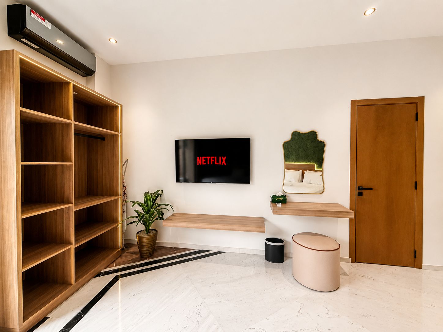 A modern hotel lobby with a large wooden bookshelf, a mounted television displaying 'NETFLIX', a small plant, and a round ottoman.