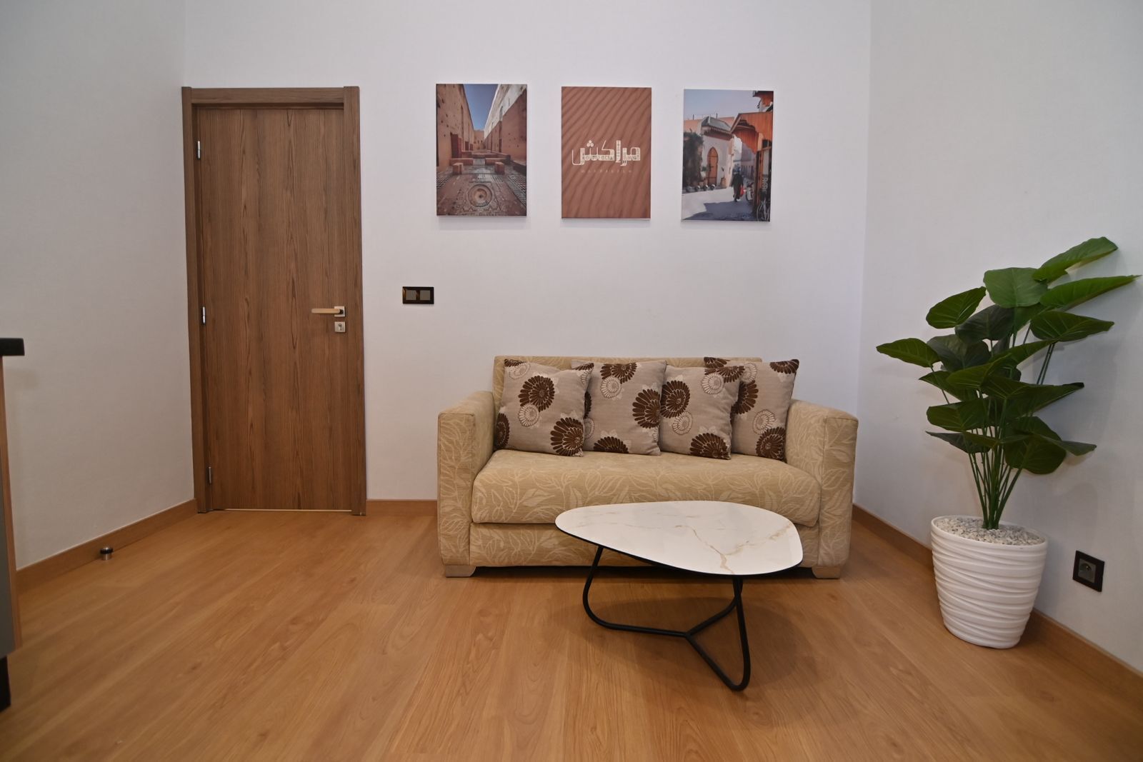 Une pièce avec une porte en bois, un canapé beige orné de coussins à motifs, une table basse blanche et une plante en pot.