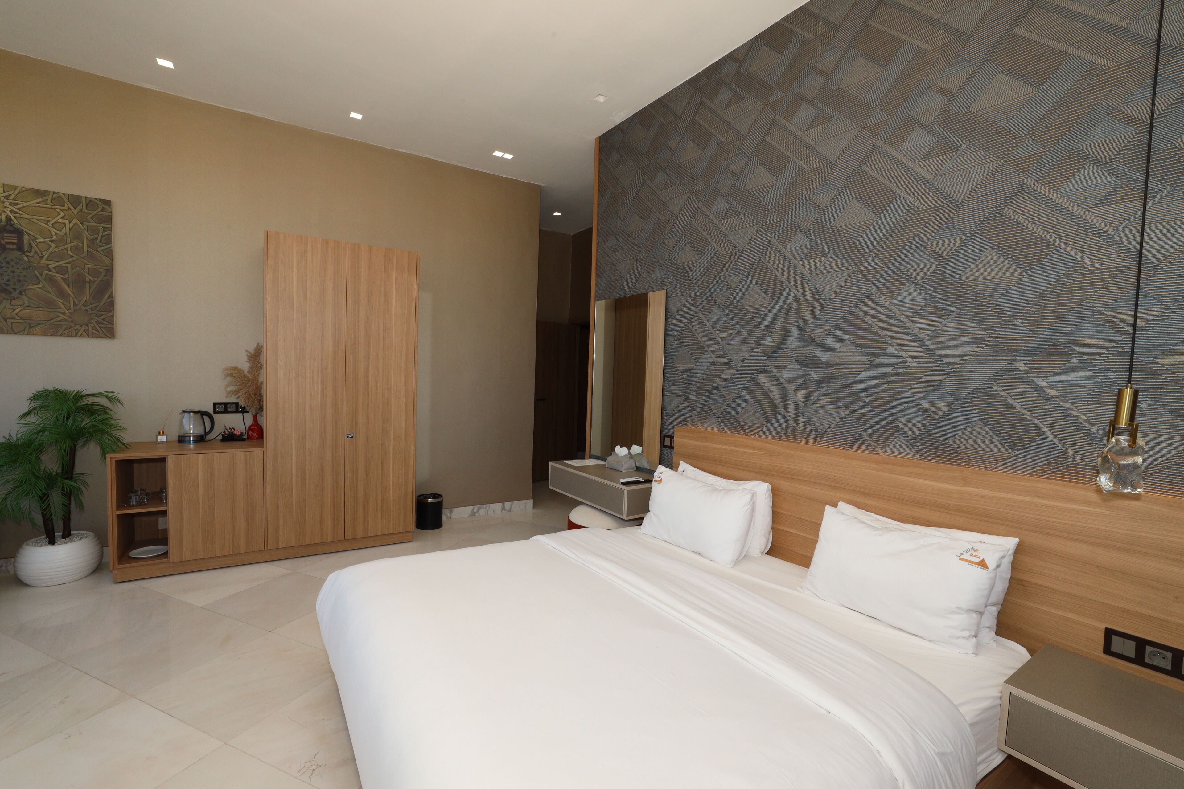 Une chambre d’hôtel moderne avec un grand lit, une armoire en bois, un petit coin salon agrémenté d’une plante, et une salle de bain visible au fond.