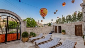A courtyard with lounge chairs, a stone wall, and hot air balloons in the background. - Soho Cappadocia (Nevşehir)