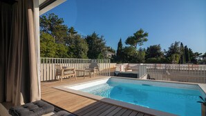 Una piscina con cubierta de madera, mobiliario exterior y vistas a árboles y edificios.