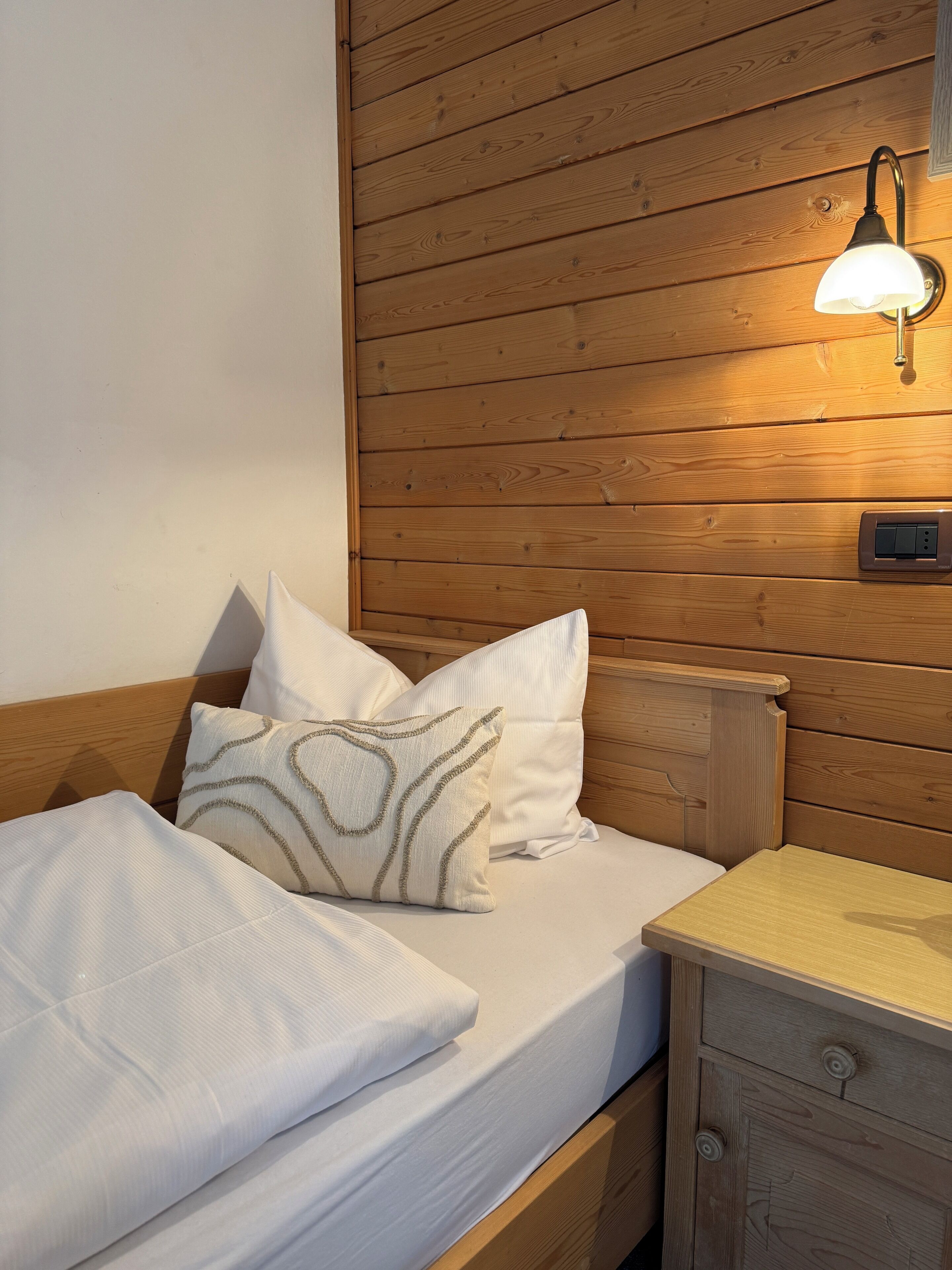 Una cama bien hecha con sábanas blancas y almohadas estampadas, un cabecero de madera, una mesita de noche con una lámpara y una cómoda pequeña.
