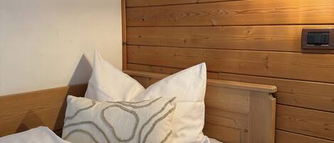 A neatly made bed with white linens and patterned pillows, a wooden headboard, a bedside table with a lamp, and a small drawer unit.