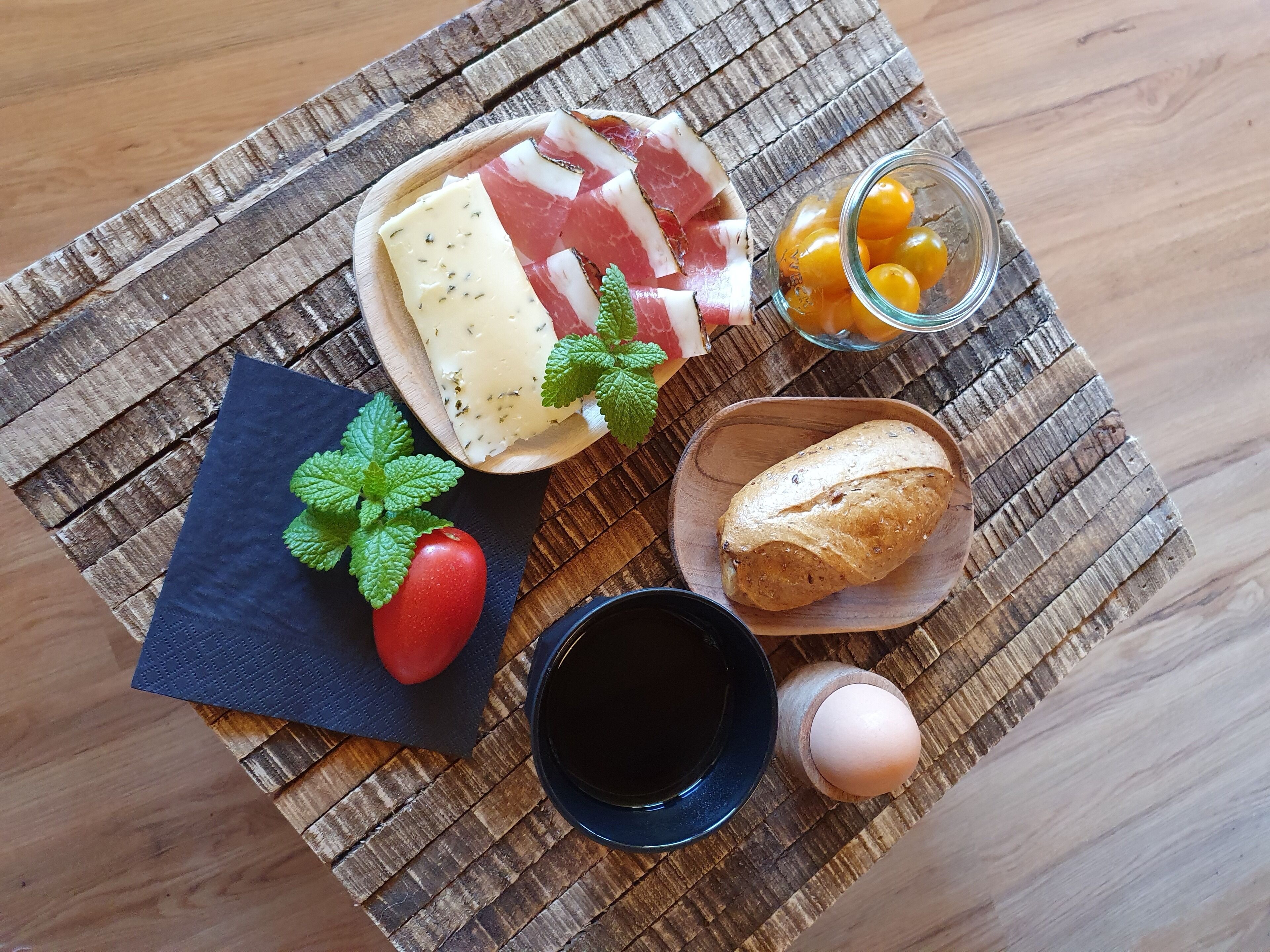 A wooden table with a variety of food items including cheese, tomatoes, a baguette, and a small glass jar.