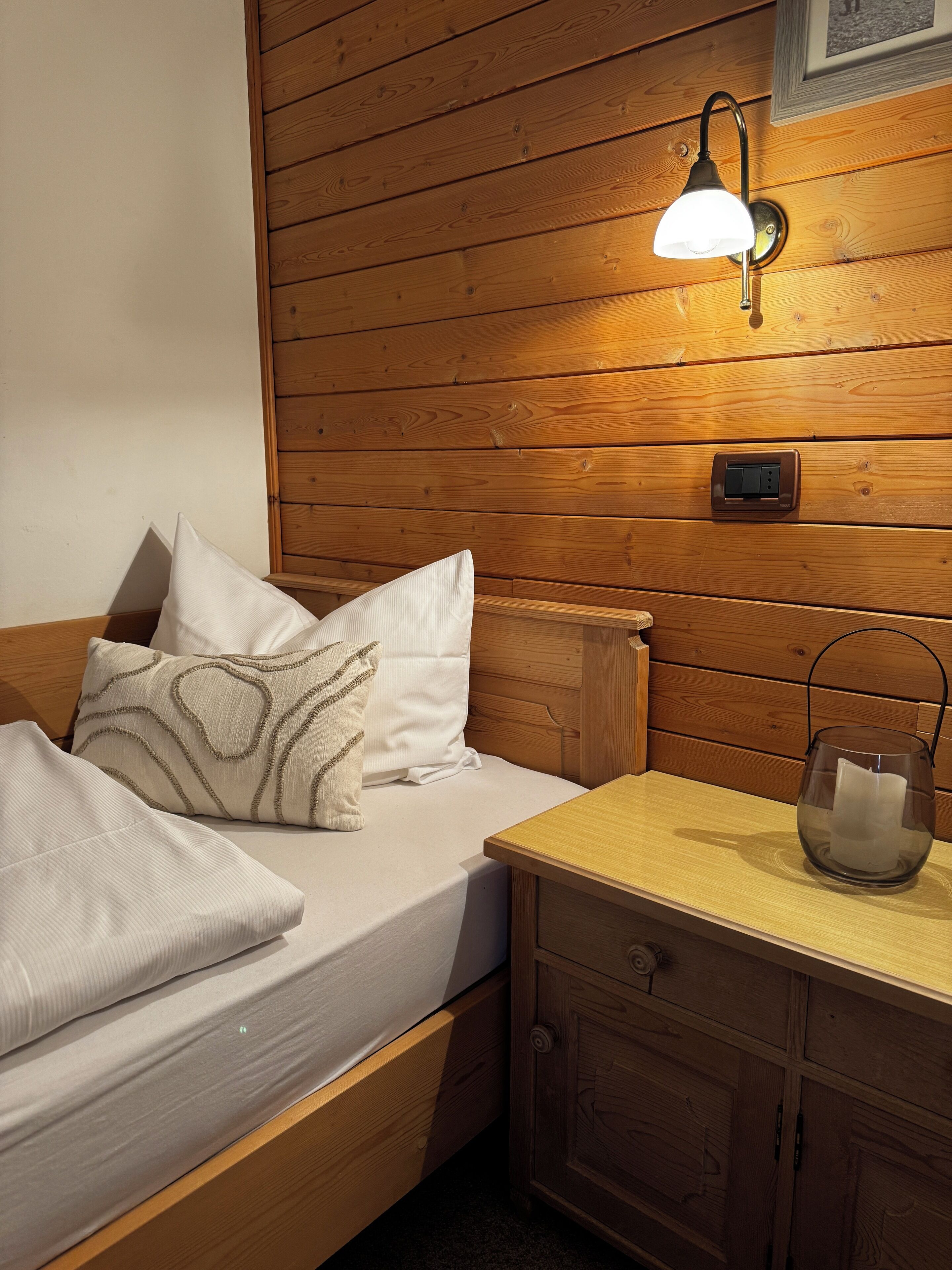 A wooden bed with white bedding and decorative pillows, a bedside table with a glass candle holder, and a wall-mounted light fixture.