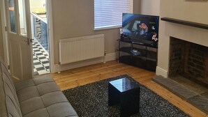 A living room with a grey sofa, a black coffee table, a fireplace, a television, and a window with blinds. - Quaint 2-bedroom house in Leicester for a cozy getaway (Leicester)