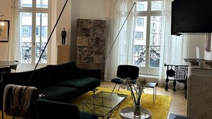 Una sala de estar moderna con un sofá verde, una mesa de centro de vidrio y un cuadro abstracto grande. Hay un televisor negro sobre un soporte, una computadora portátil sobre una mesa y una silla con un tapete amarillo.