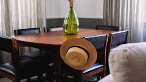 Una mesa de comedor con sillas, un florero verde con flores secas y un sombrero de paja sobre una silla.