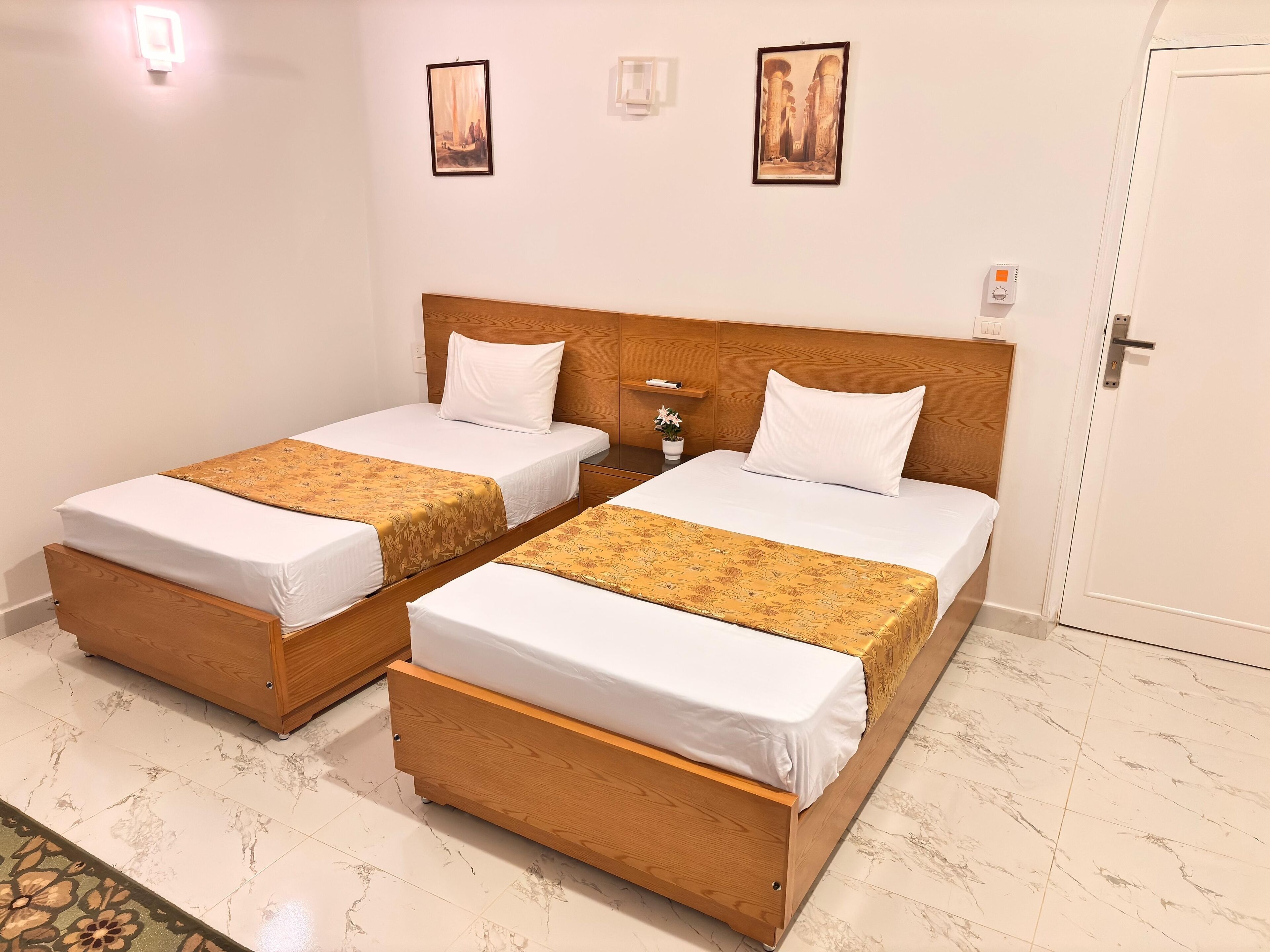 A hotel room with two wooden beds, white bedding, a small bedside table with a lamp, and a wall-mounted thermostat.