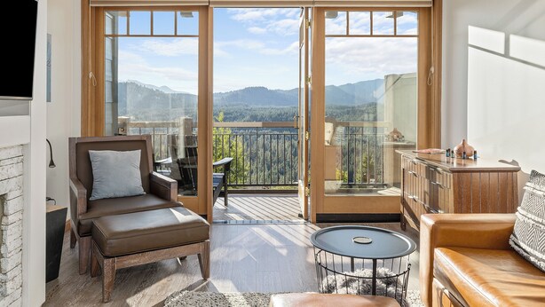 A modern living room with a brown leather sofa, a small round table, and a wooden cabinet. A large sliding glass door leads to a balcony with a view of mountains and trees. - Tranquil Suncadia Lodge with Fireplace, Mountain Views and Pool Hot Tub (Cle Elum)