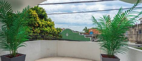 A balcony with two potted plants, a tiled floor, and a view of a green building and a blue sky.