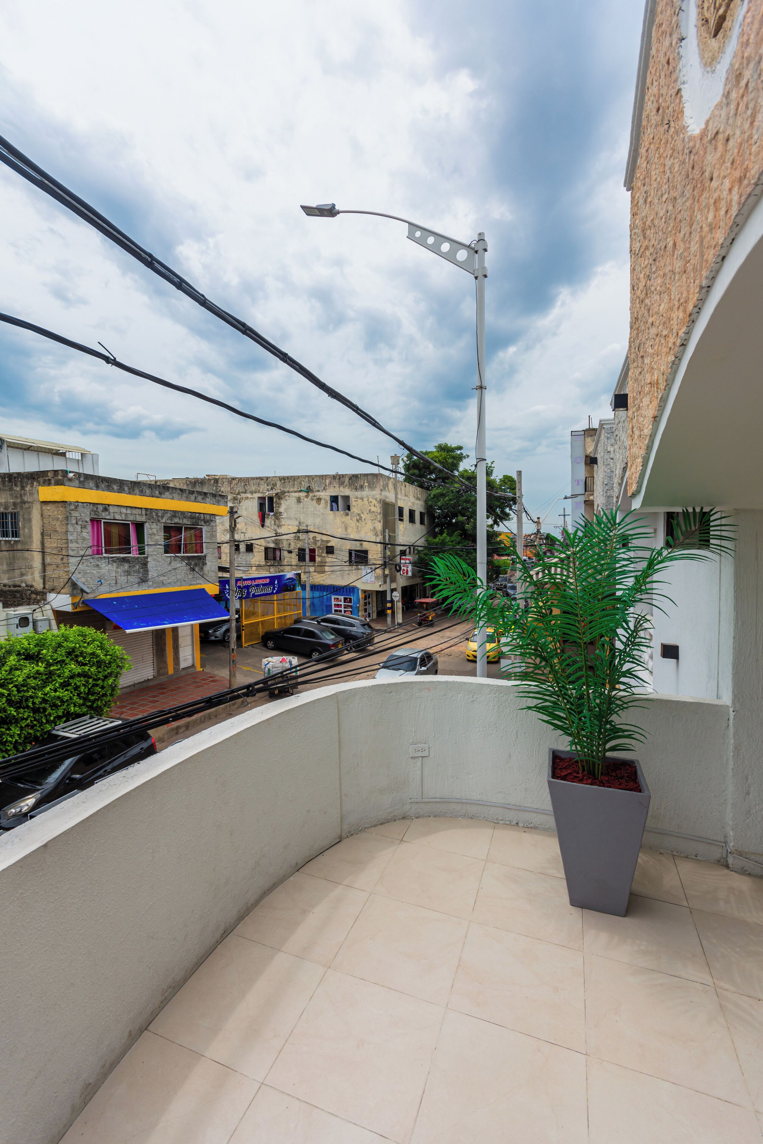 A balcony with a potted plant, a tiled floor, and a view of a street with buildings and a tram.