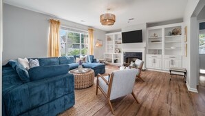 Ein Wohnzimmer mit einem blauen Sofa, einem Korbtisch und einem weißen Sessel. Ein Kamin ist vorhanden, darüber ein Fernseher, daneben ein Bücherregal mit Dekoration.