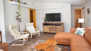 Un salon moderne avec une table basse en bois, deux fauteuils blancs, un canapé marron et un téléviseur à écran plat posé sur un meuble en bois.