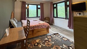 A bedroom with a bed, a desk, a chair, a TV, and large windows. - Holidays in Haven Hospitality  (Manali)
