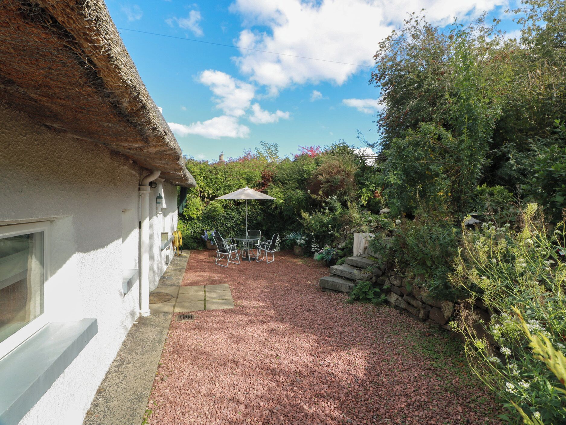 Ein Garten mit einer Terrassentischgruppe, umgeben von dichtem Grün und einer Steinmauer.