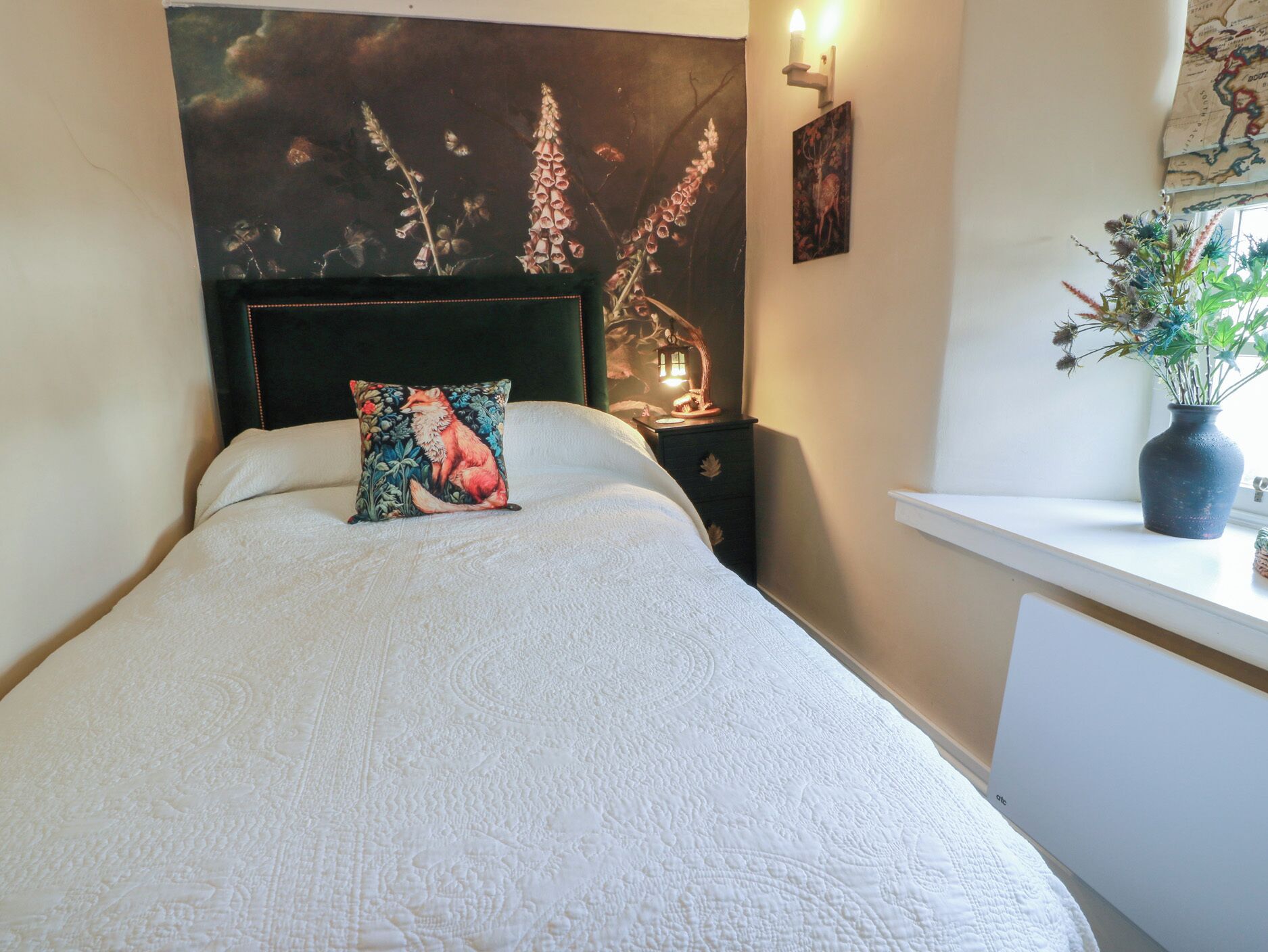 A neatly made bed with a decorative pillow, a headboard with a floral tapestry, a bedside table with a lamp, and a window with a view of the outdoors.