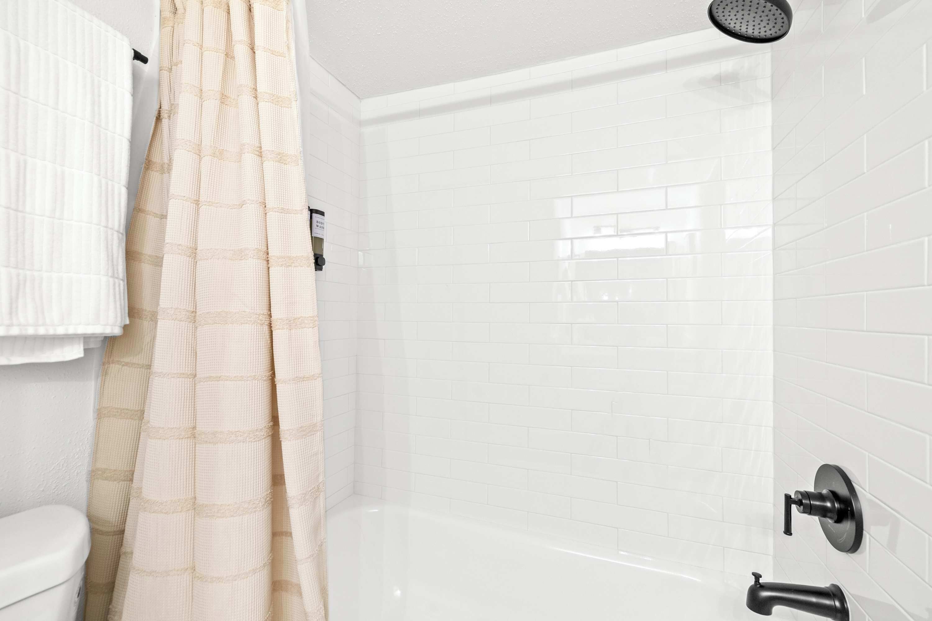 Un baño con ducha de azulejos blancos, cortina de ducha color beige y un inodoro blanco.