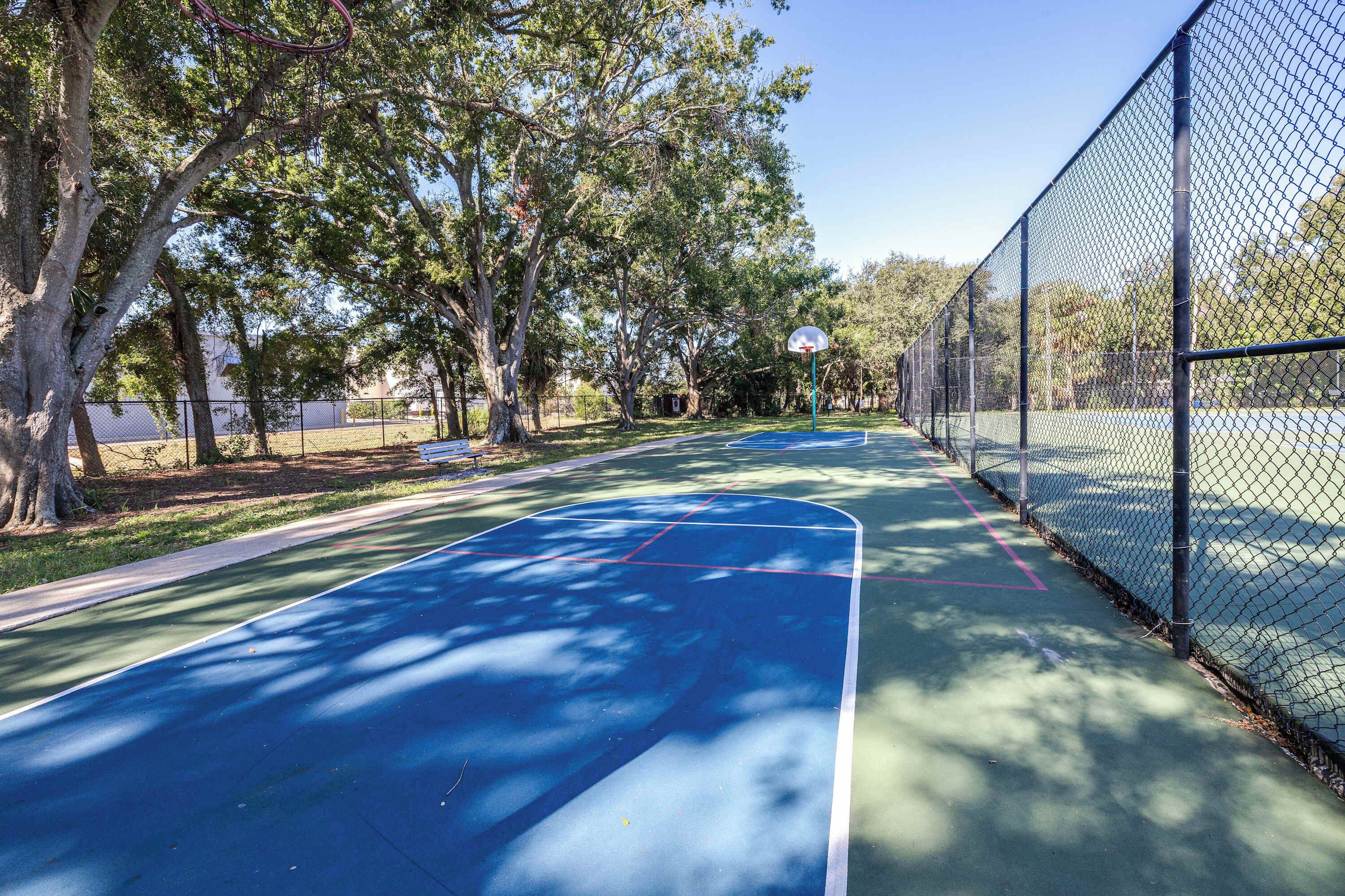 Ein Basketballplatz im Freien mit einem Drahtzaun, Bäumen und blauem Himmel.