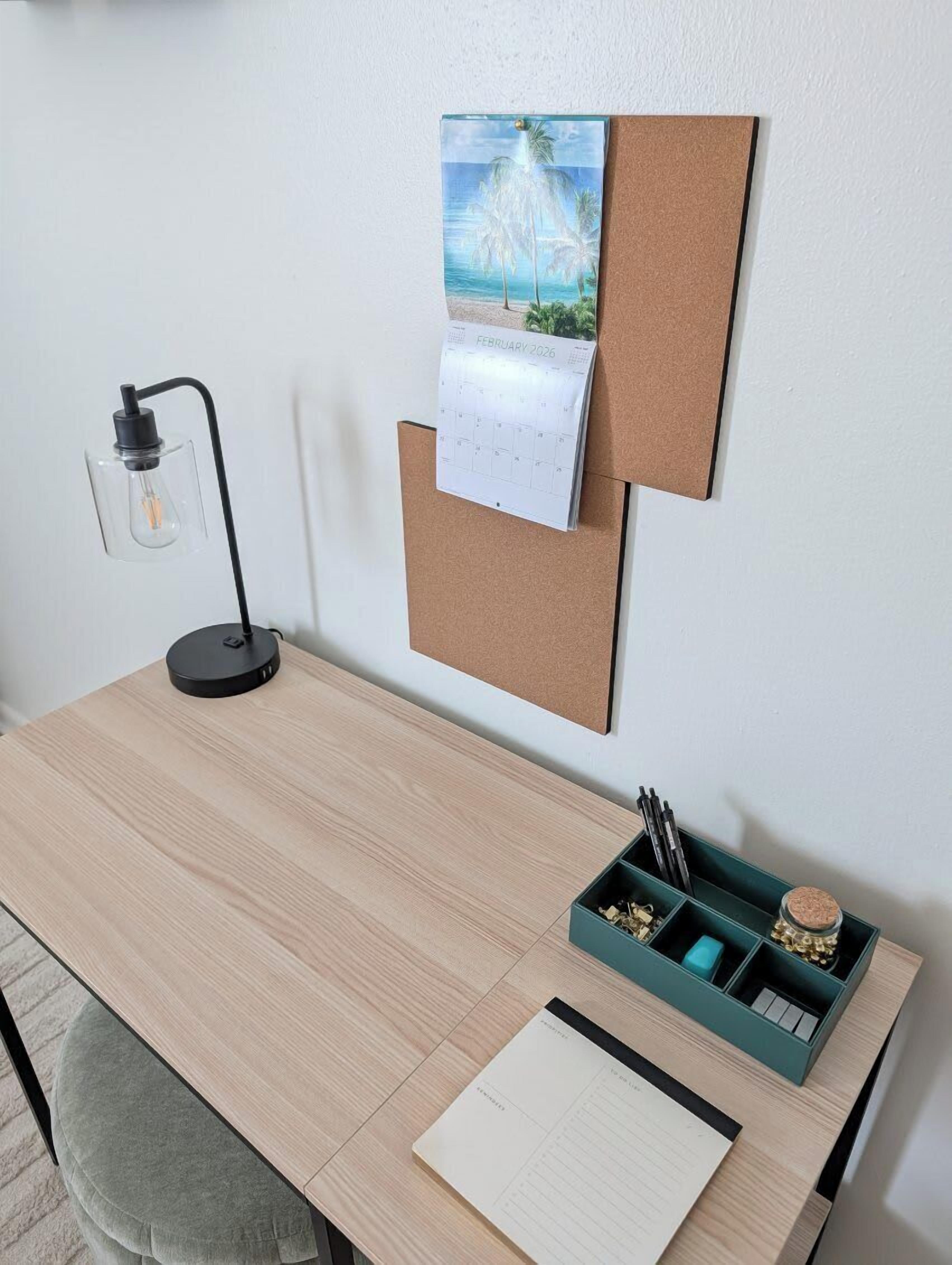 Un escritorio de madera con un calendario de corcho, una lámpara de escritorio, un bloc de notas y un portalápices con instrumentos de escritura.