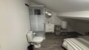 Habitació petita i senzilla amb un llit individual, una taula de nit, un mirall, un vàter i un lavabo.