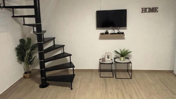 Un saló modern amb una escala espirala negra, un televisor de pantalla plana muntat a la paret i dues taules laterals amb objectes decoratius.
