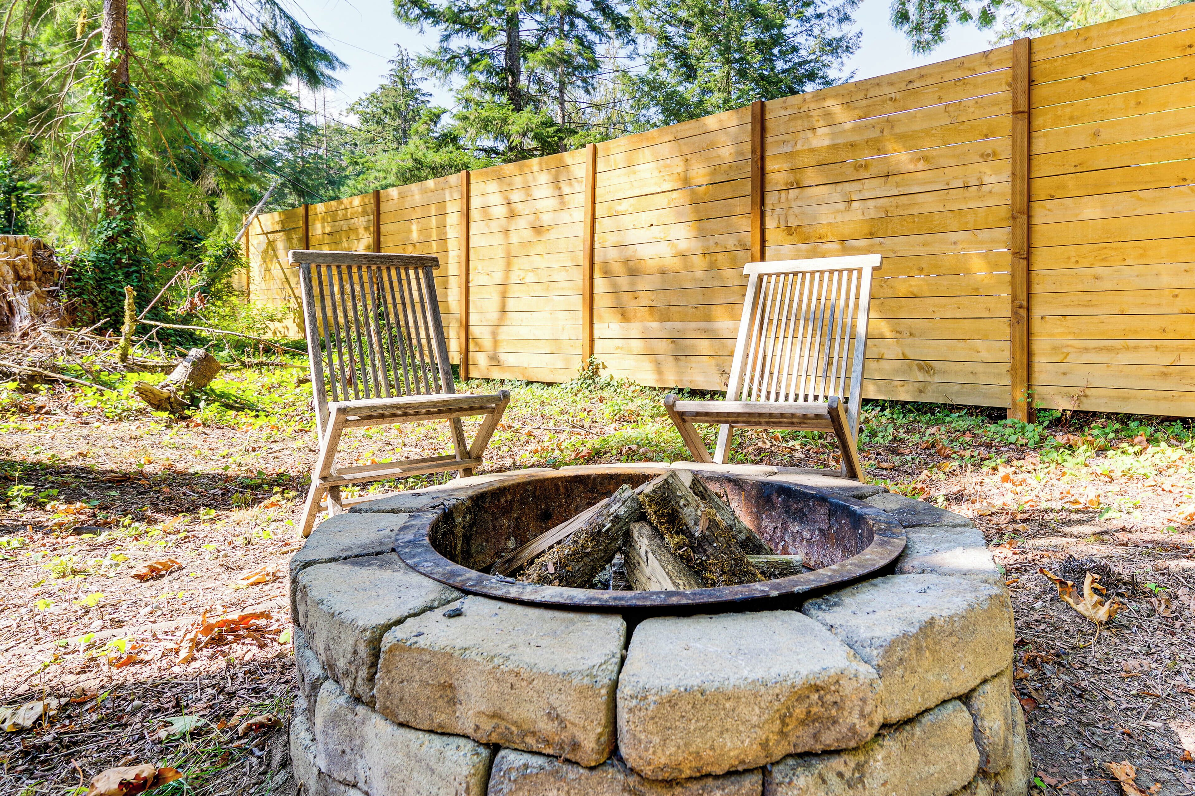A stone fire pit with two wooden chairs, surrounded by a wooden fence and trees.